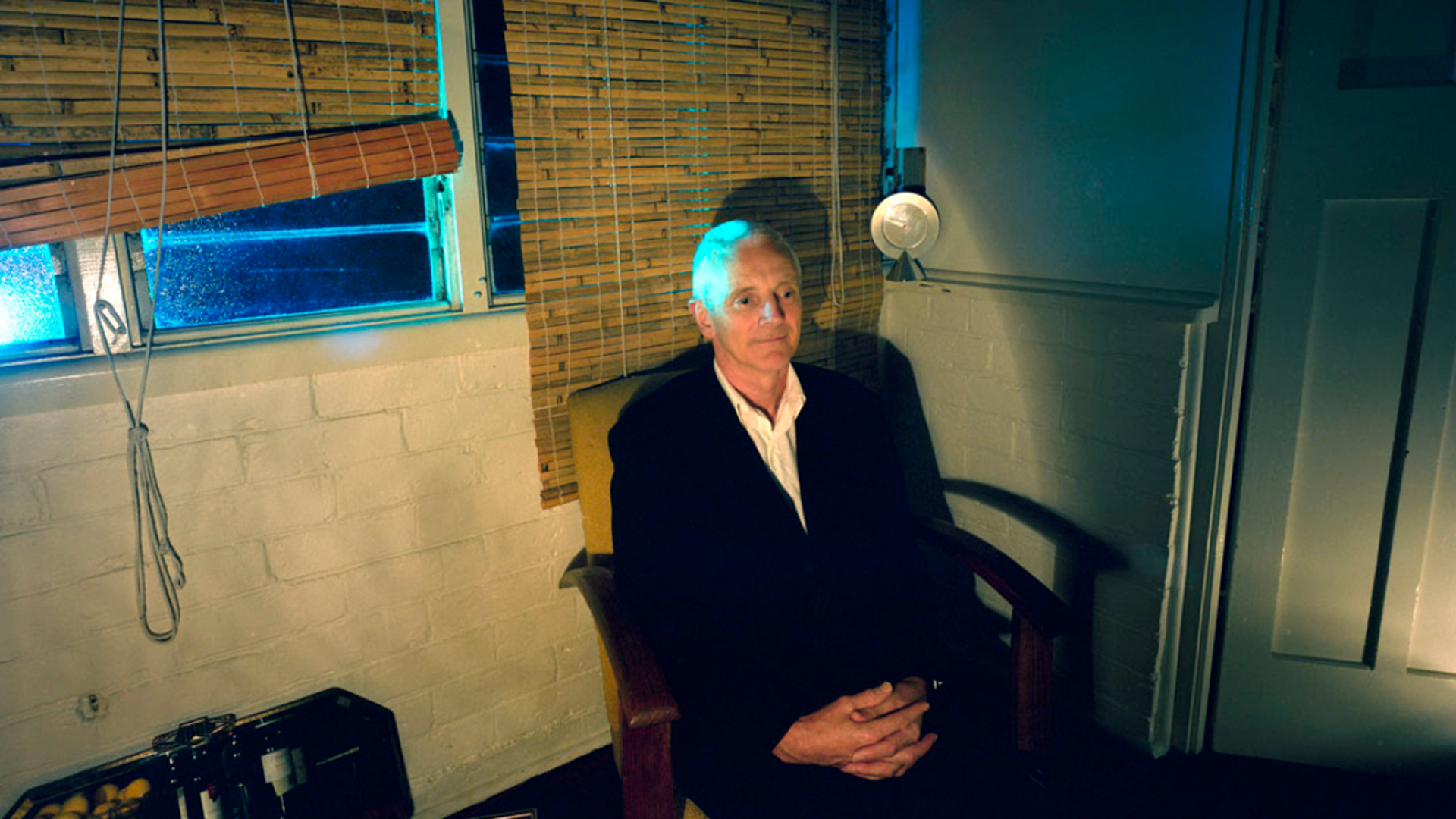 ID: A bald man wearing a suit is sitting in an armchair. The room around him is dark, dimly lit by a lamp outside the frame.