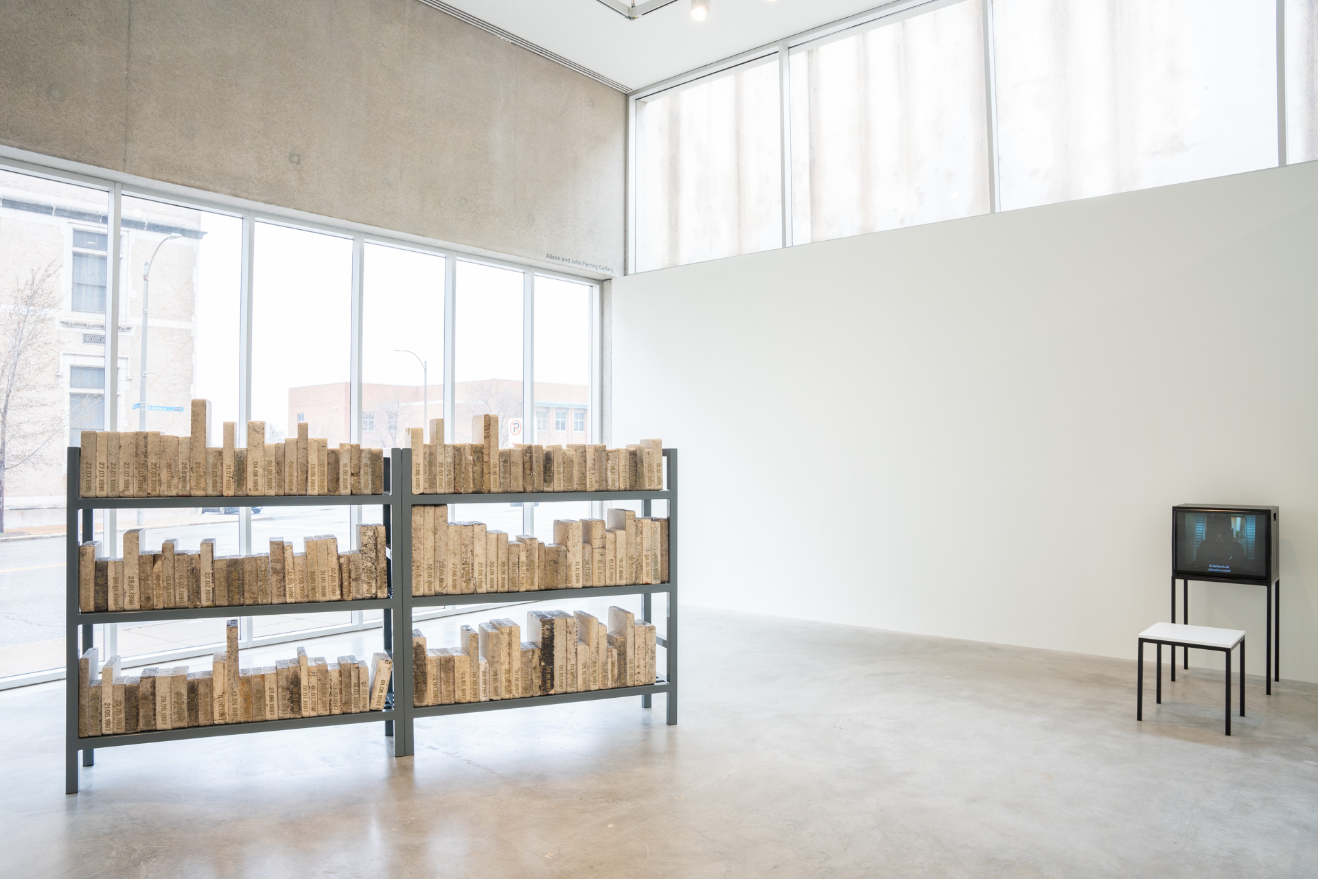 A sparse, white walled museum gallery with lots of natural light coming in through two large windows. In the center of the floor is what seems like two sections of old library texts, three shelves tall each. The colors of the items on the shelves are washed out to whites, light beiges, and light browns. There is a small CRT TV against an adjacent wall with a small white stool in front of it.