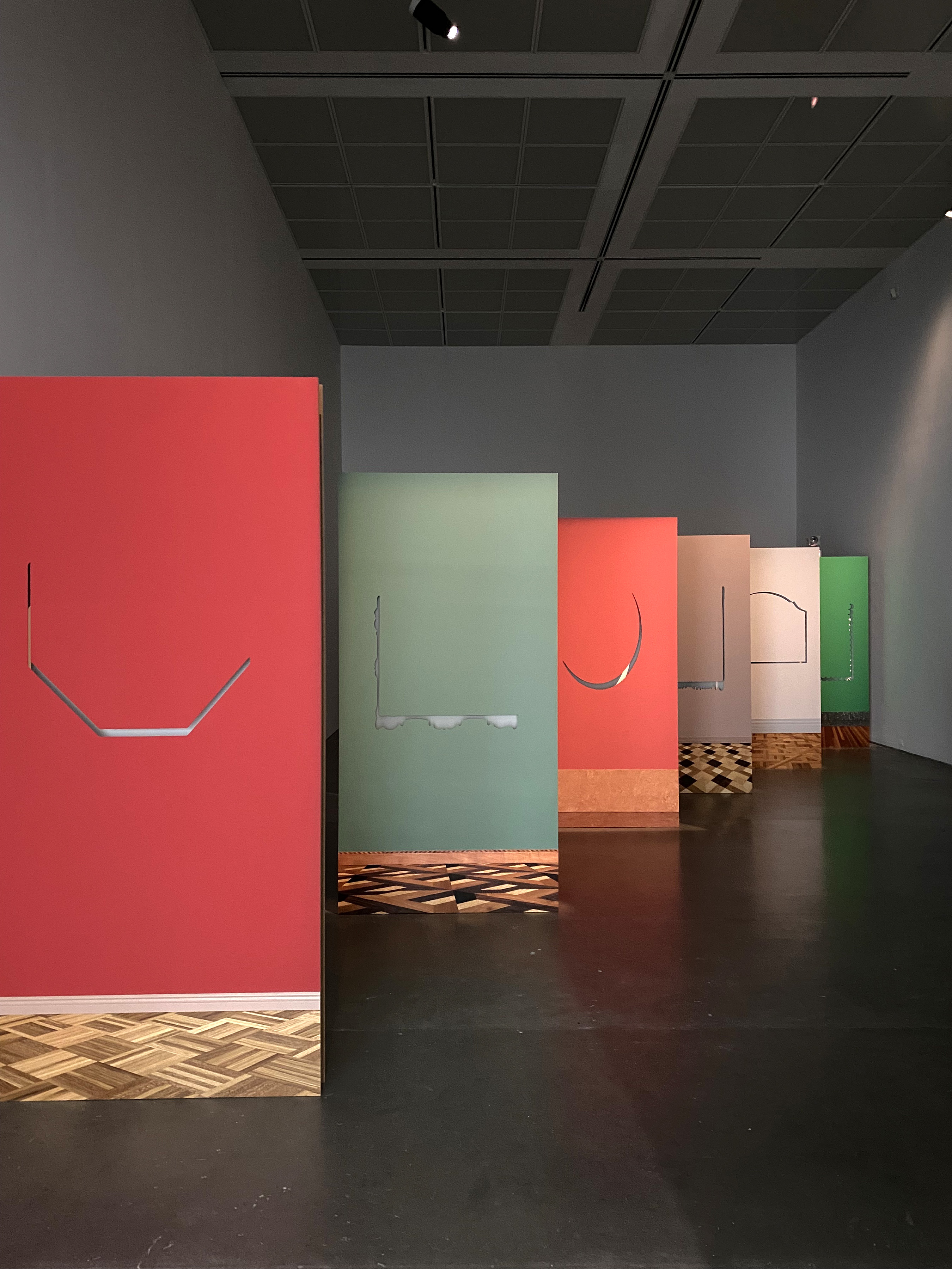 A dimly lit art gallery with white walls and gray floors has 6 large paintings or images free standing on the floor and across the room in a diagonal line, receding from view. They are mostly of flat color fields with thin glyphic marks that resemble letters in their center.