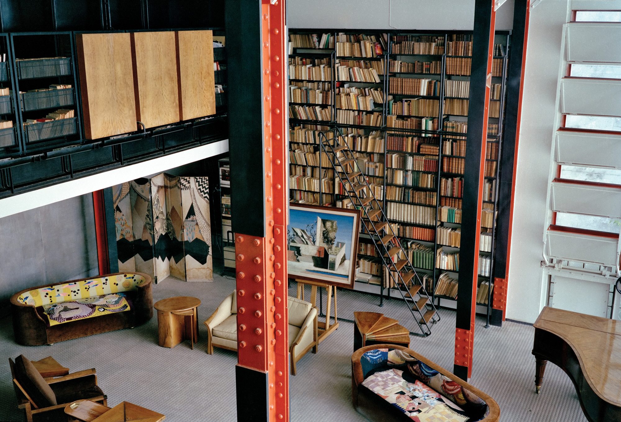Maision de Verre’s great living room taken by the second floor. The room has a grand book case filled with books and gorgeous furniture. There is black and red industrial beams supporting the open concept.