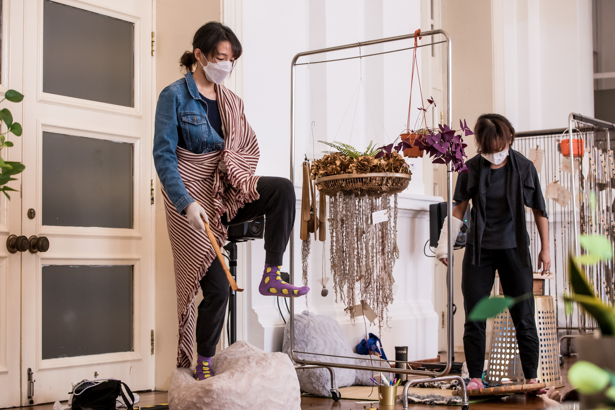 IMAGE DESCRIPTION: Another photo from our workshop; a different masked participant in a blue denim jacket has a red-and-white striped blanket draped over her left shoulder and right hip, and is balancing on one leg on a fuzzy bean bag. She is also holding a backscratcher as if about to scratch the soles of her own feet. Separated by a hospital frame from which real and artificial plants hang, another masked person looks down at a round bamboo tray, which she has sandwiched between her feet.