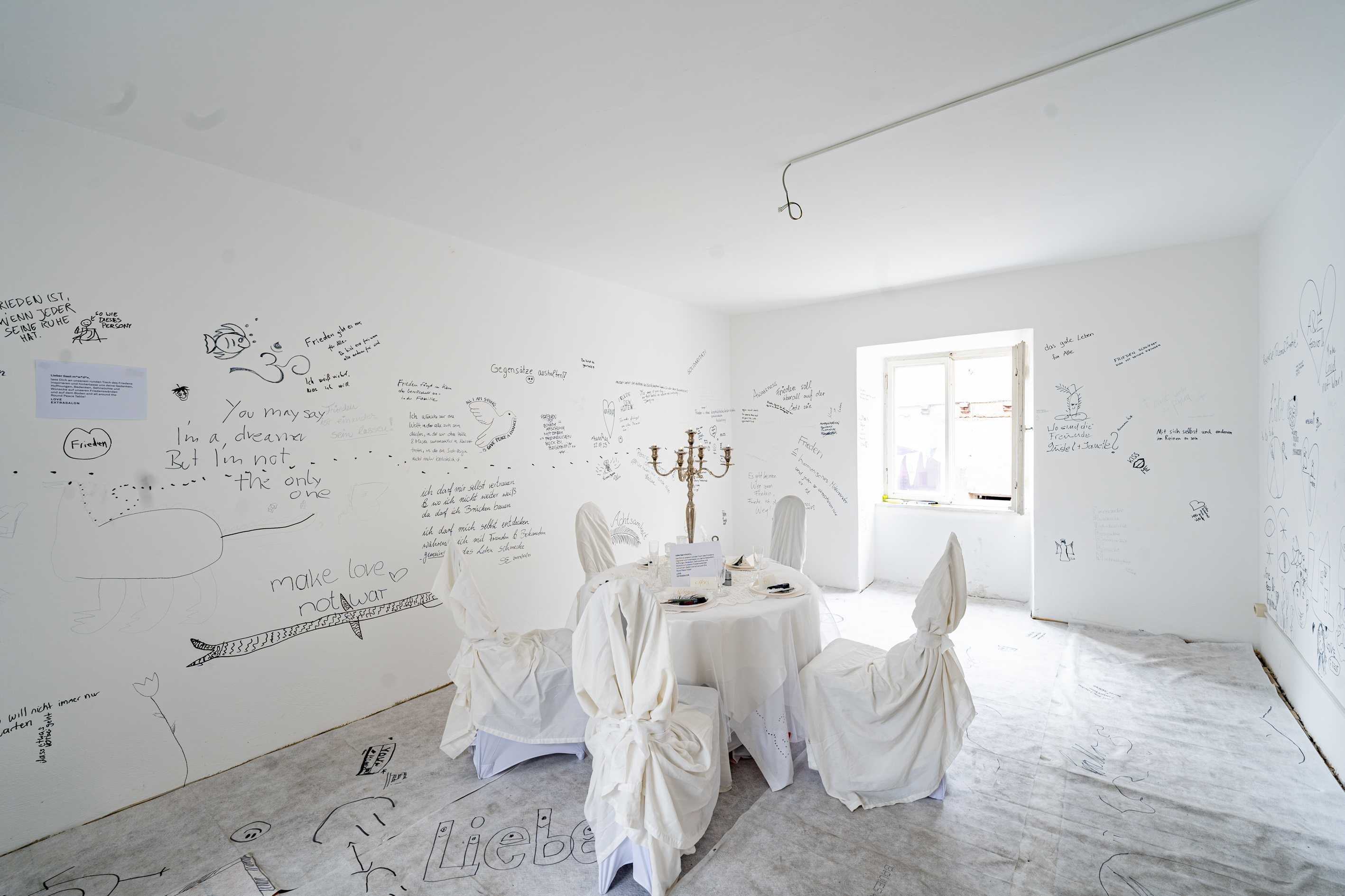 Bild 1 Tischlein deck dich: Ein wei&szlig;er Raum mit zwei Fenstern rechts und links. In der Mitte ein Tisch mit wei&szlig;er Tischdecke und 5 mit wei&szlig;em Stoff verh&uuml;llte St&uuml;hle. Die Besuchenden k&ouml;nnen W&auml;nde und Boden beschreiben.