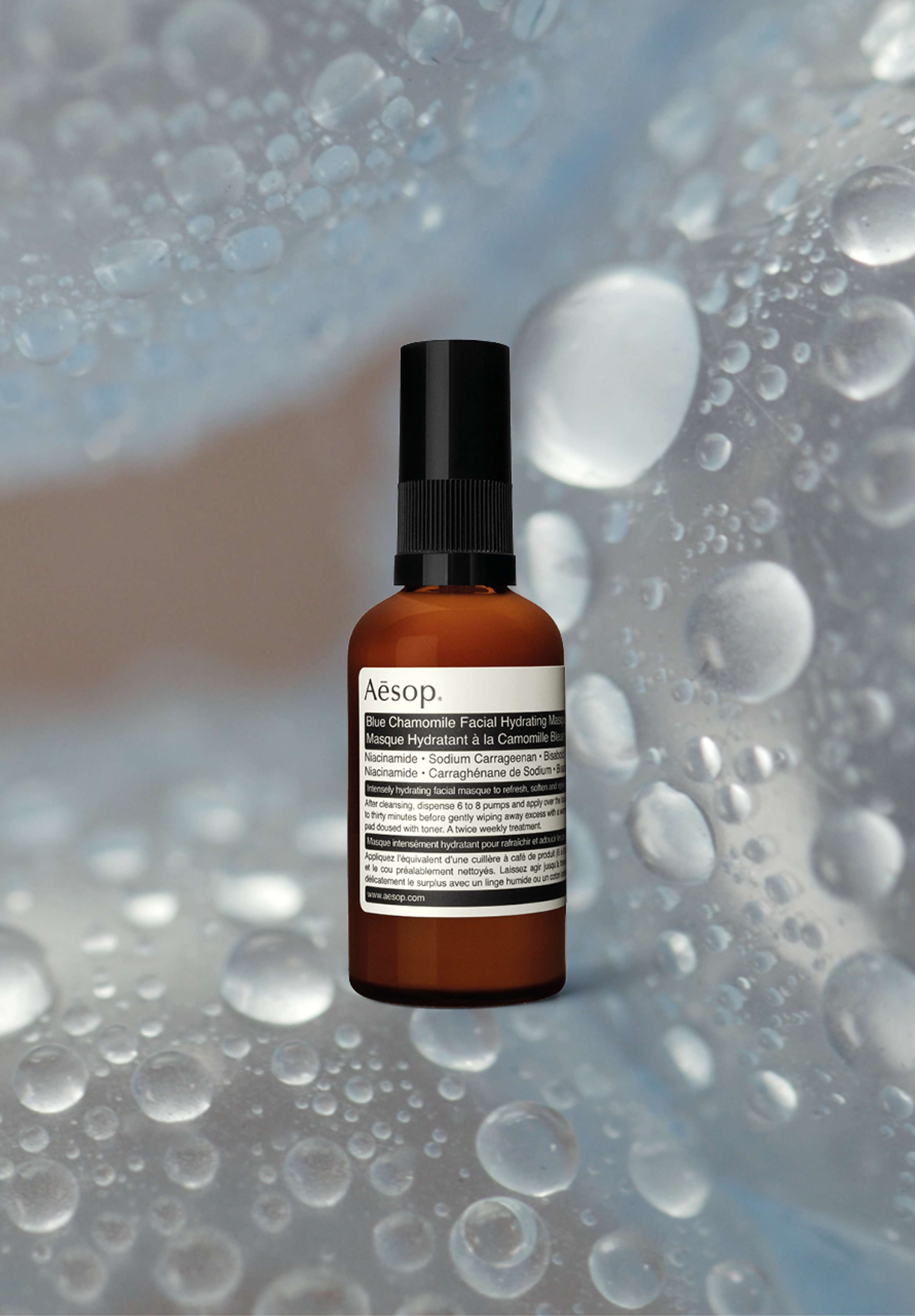 A bottle of Aesop's Blue Chamomile Facial Hydrating Masque is overlaid on top of macro photography of water droplets on blue spheres.