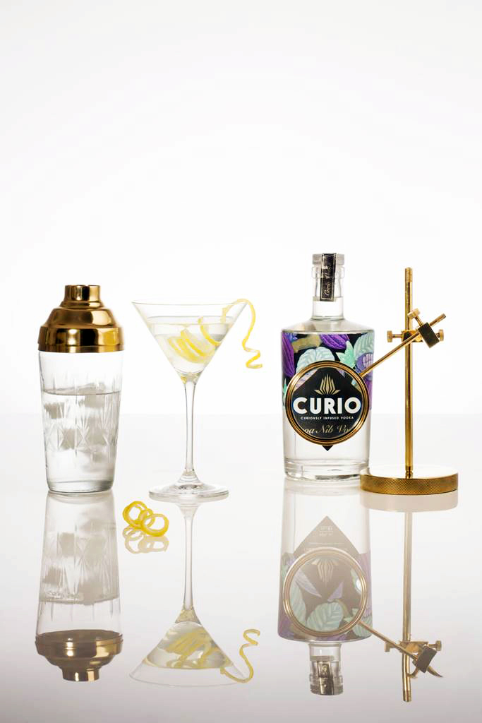 Minimal composition of cocktail shaker, cocktail glass, bottle of Curio and magnifying equipment on a reflective surface.