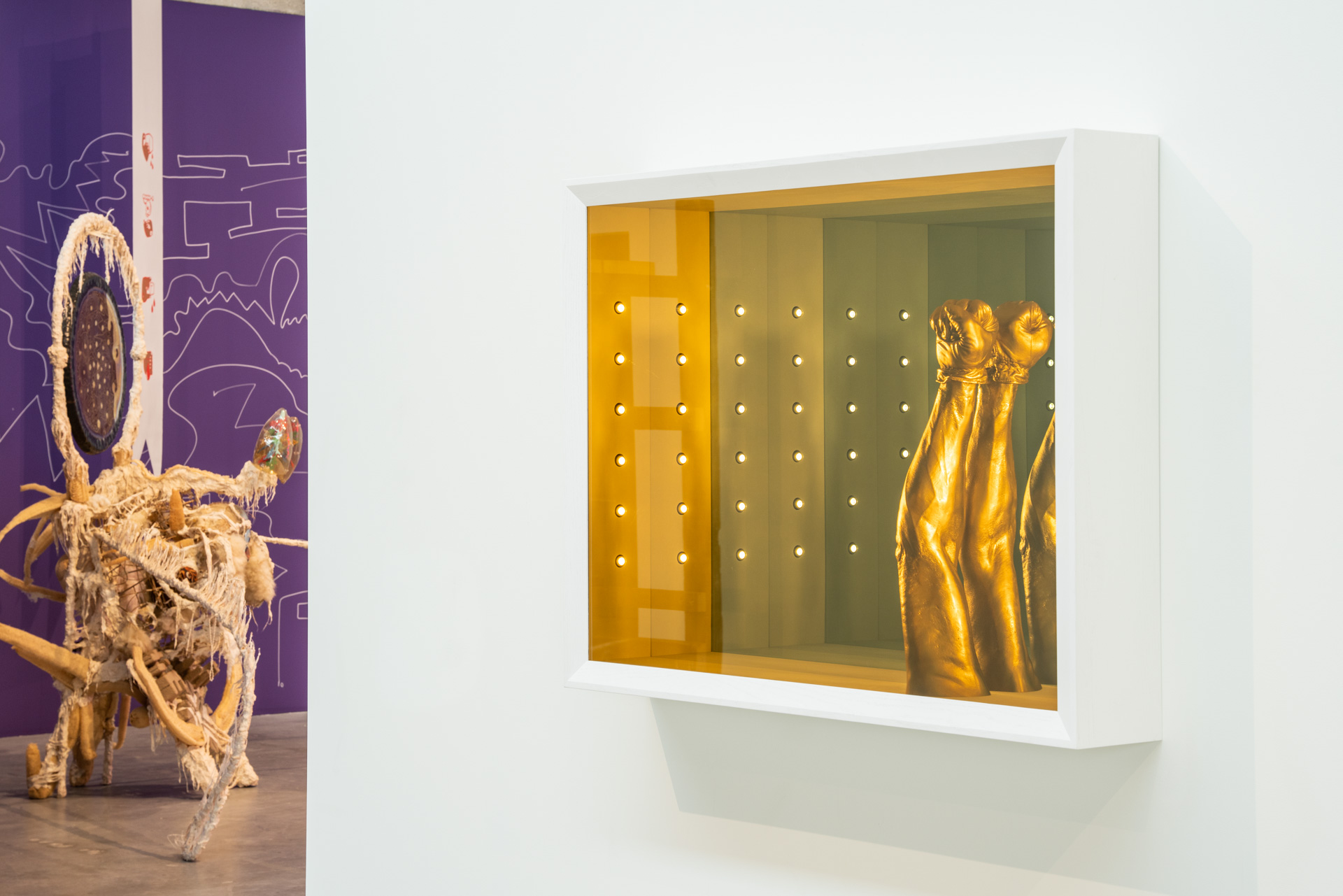 A wall mounted abstract sculpture on a white wall in the form of a shallow shadow box with yellow glass in the front, and a golden fist, pointing upward, inside.