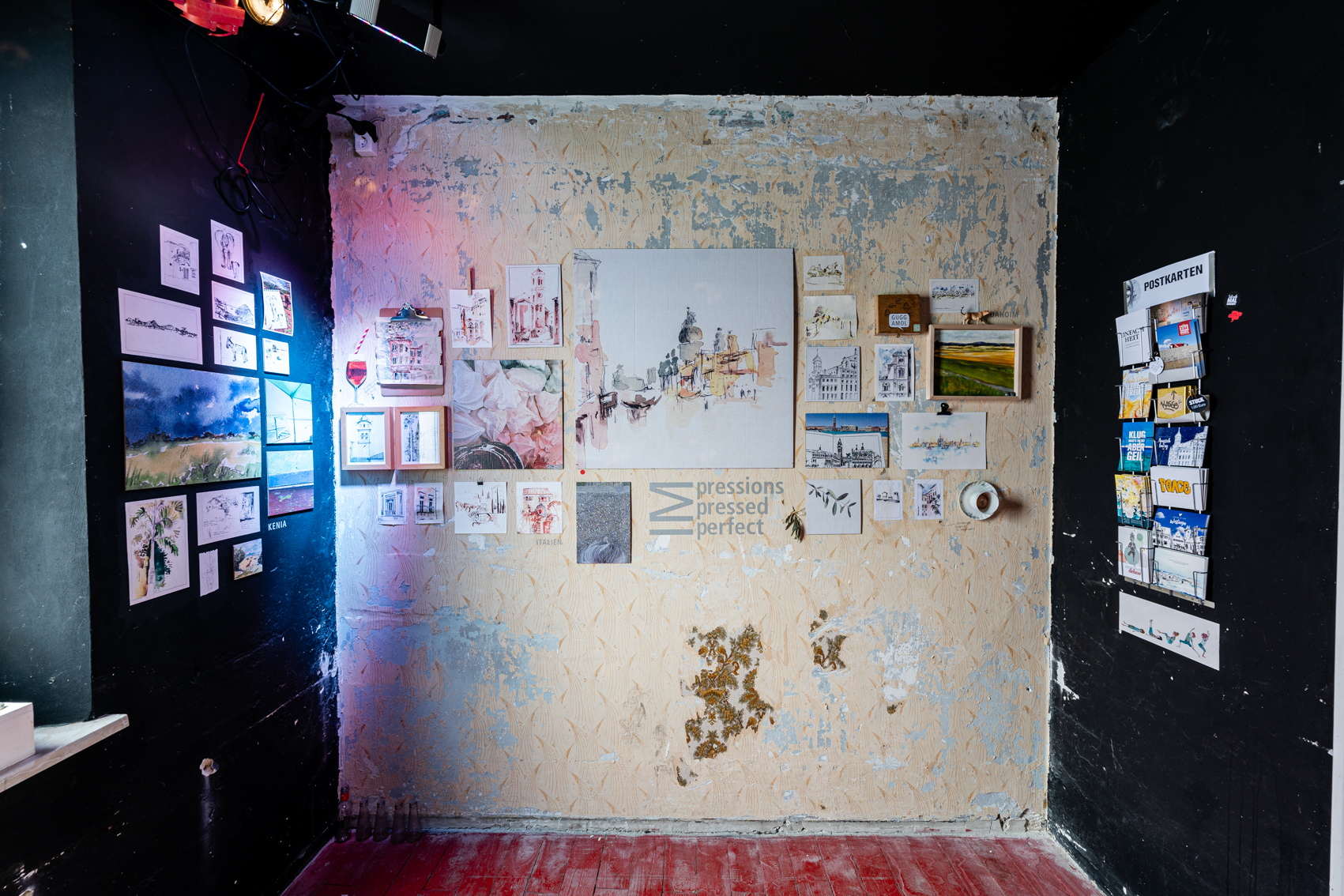 Bild 1 zu IMpressions | IMpressed | IMperfect: Quadratischer Raum. Decke und Seitenwände sind schwarz gestrichen. Die Wand frontal ist beige, die Tapete blättert teils ab. An den Wänden hängen verschiedene Aquarelle und Gegenstände. Vorwiegende Motive sind Städte und Landschaften.