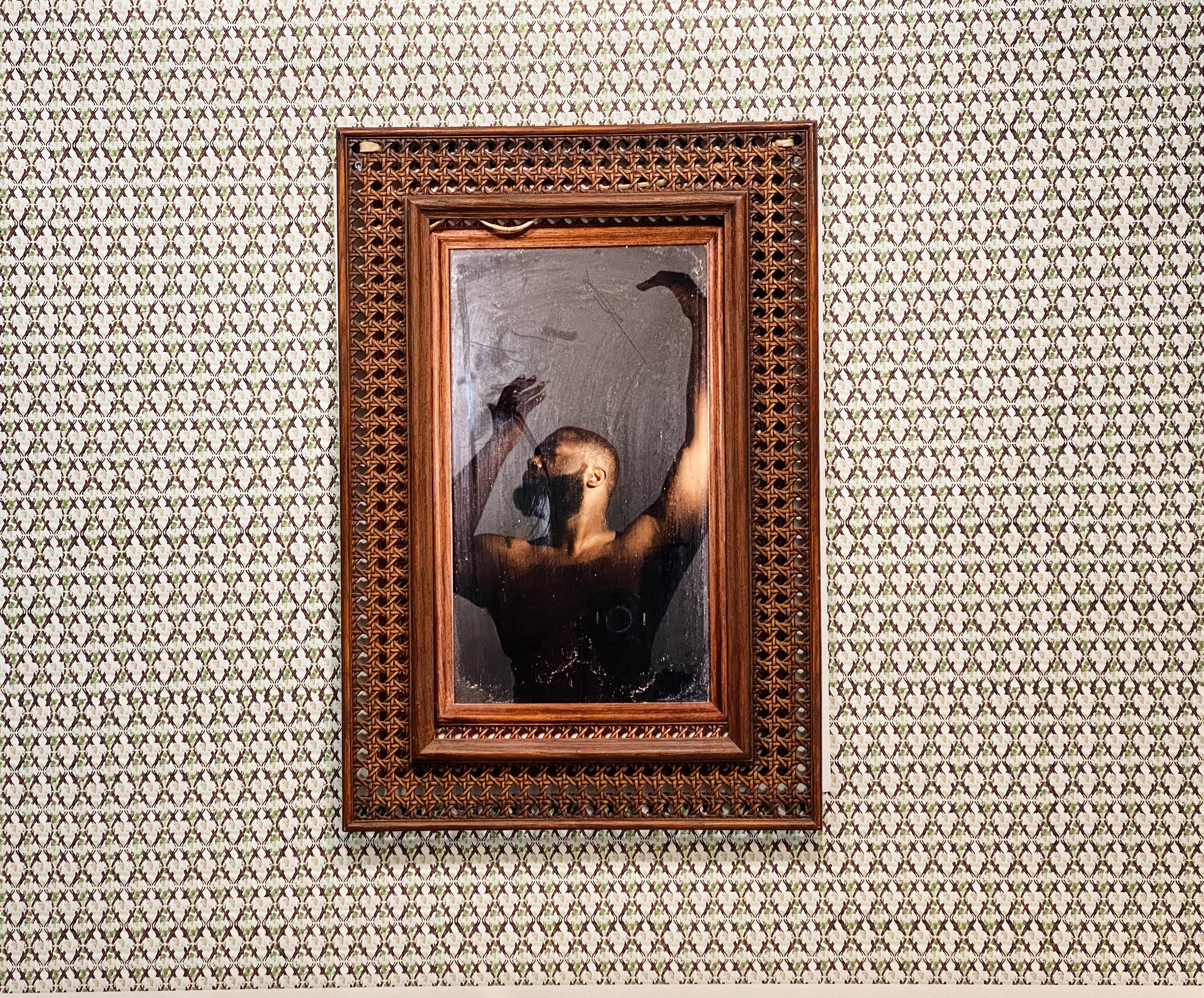 A painting from the waist up of a nude figure with medium dark skin and a beard, who has both hands gracefully raised above their head that has been framed with what seems like wicker furniture material against a dense black and white pattern wallpaper.