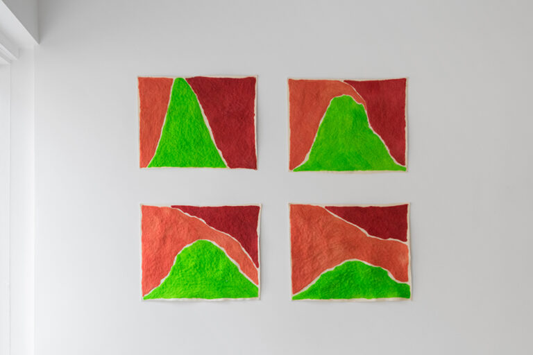 Pictured: Four square pieces of hand dyed wool mounted on a white wall, dyed red, green and orange in indistinct sections 