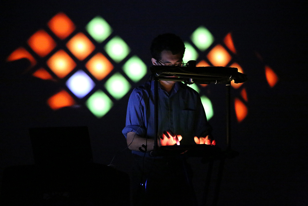 A half body shot of Daniel Corral performing in the dark with video art in the background.