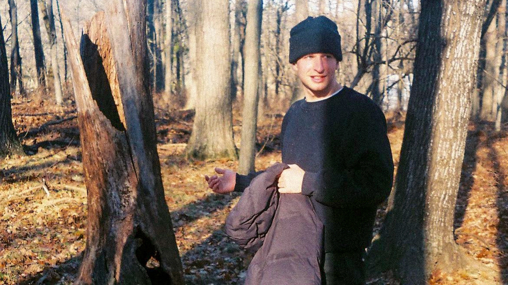 a man in the woods holding a coat with a black beanie