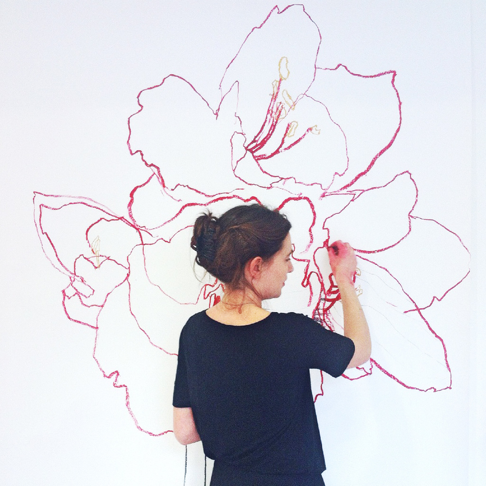 The artist Chloe Briggs is drawing a flower in red chalk on a white wall