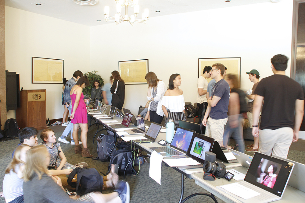 A crowded room where people chat and view video artworks displayed on a long table.