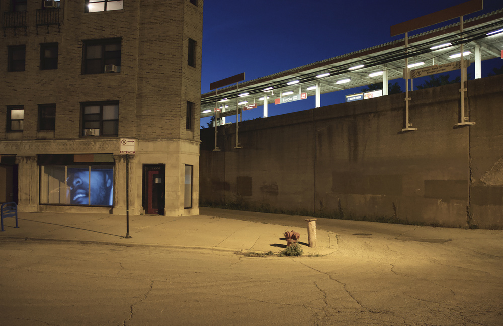 Street view of projected moving image work by Paige Taul. Through storefront windows, black and white video shows a face from the bridge of the nose to the chin, facing left. The projected image fills the window space.