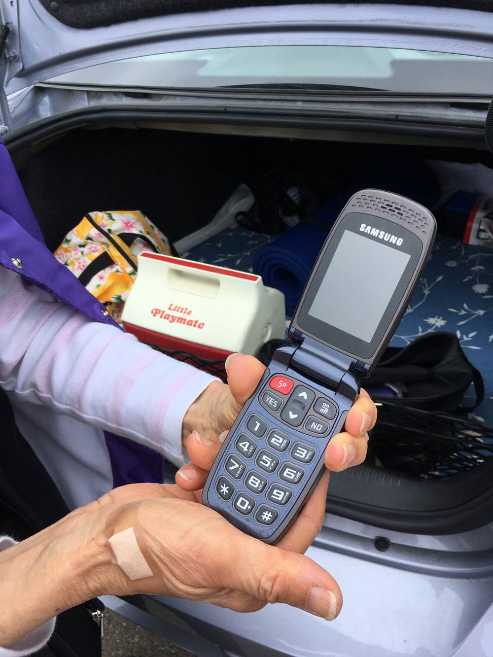 A pair of hands holds an open Samsung flip phone, in front of an open car trunk