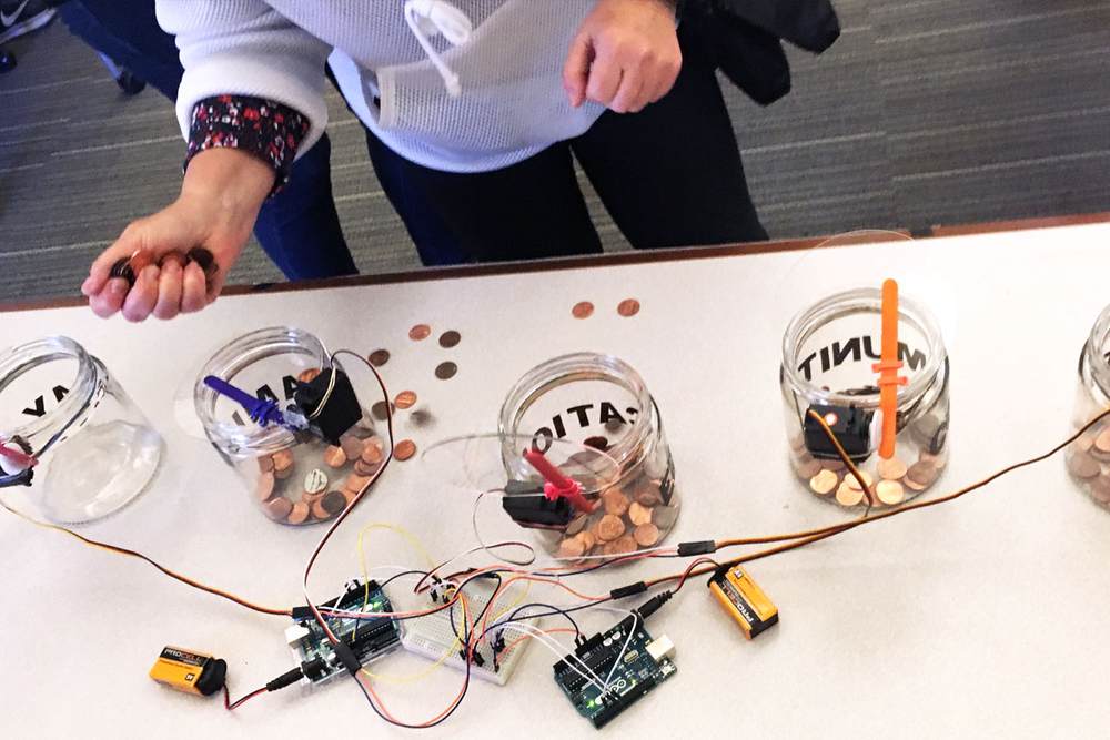 A person holds a fistful of pennies, holding it over a jar. Next to it are other jars, and behind the jars are two Arduinos powered by 9-volt batteries.