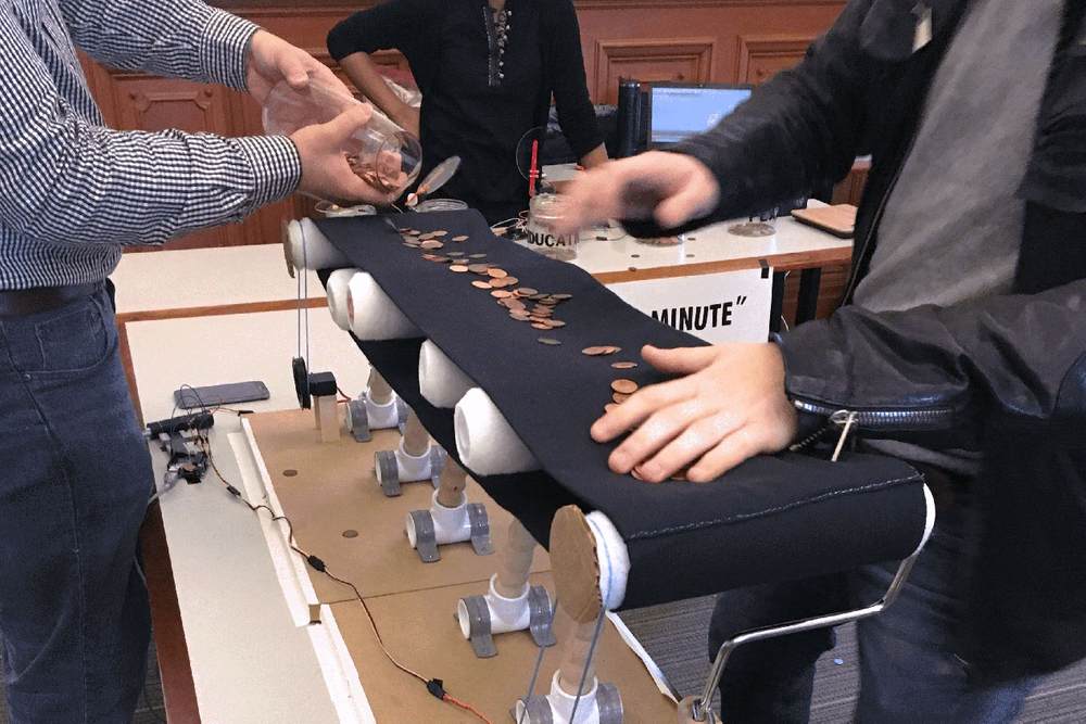 A person empties coins onto a handmade conveyor belt, as another person uses both hands to grab coins