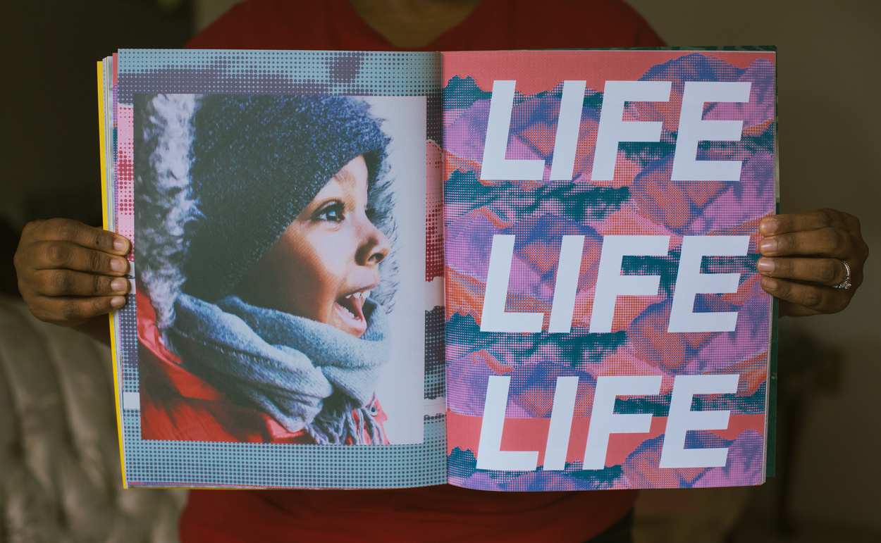 zine held open by african american hands to left hand page page showing profile image of smiling african american boy in knit cap and hooded parka and right hand page showing stylized red and violet background with LIFE LIFE LIFE printed over in block italicized letters.