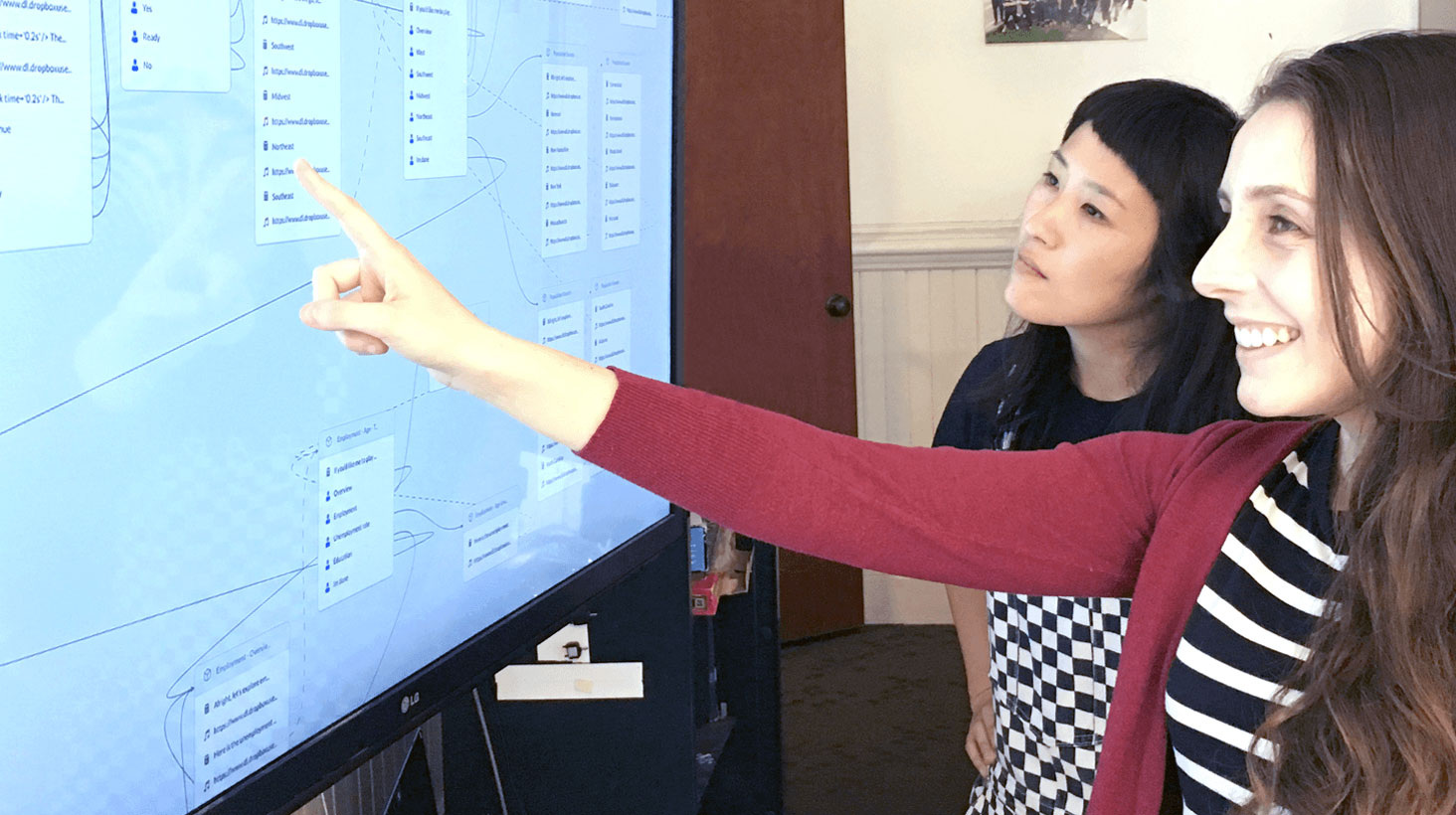 Two young women stand looking at a large screen. One is smiling and pointing