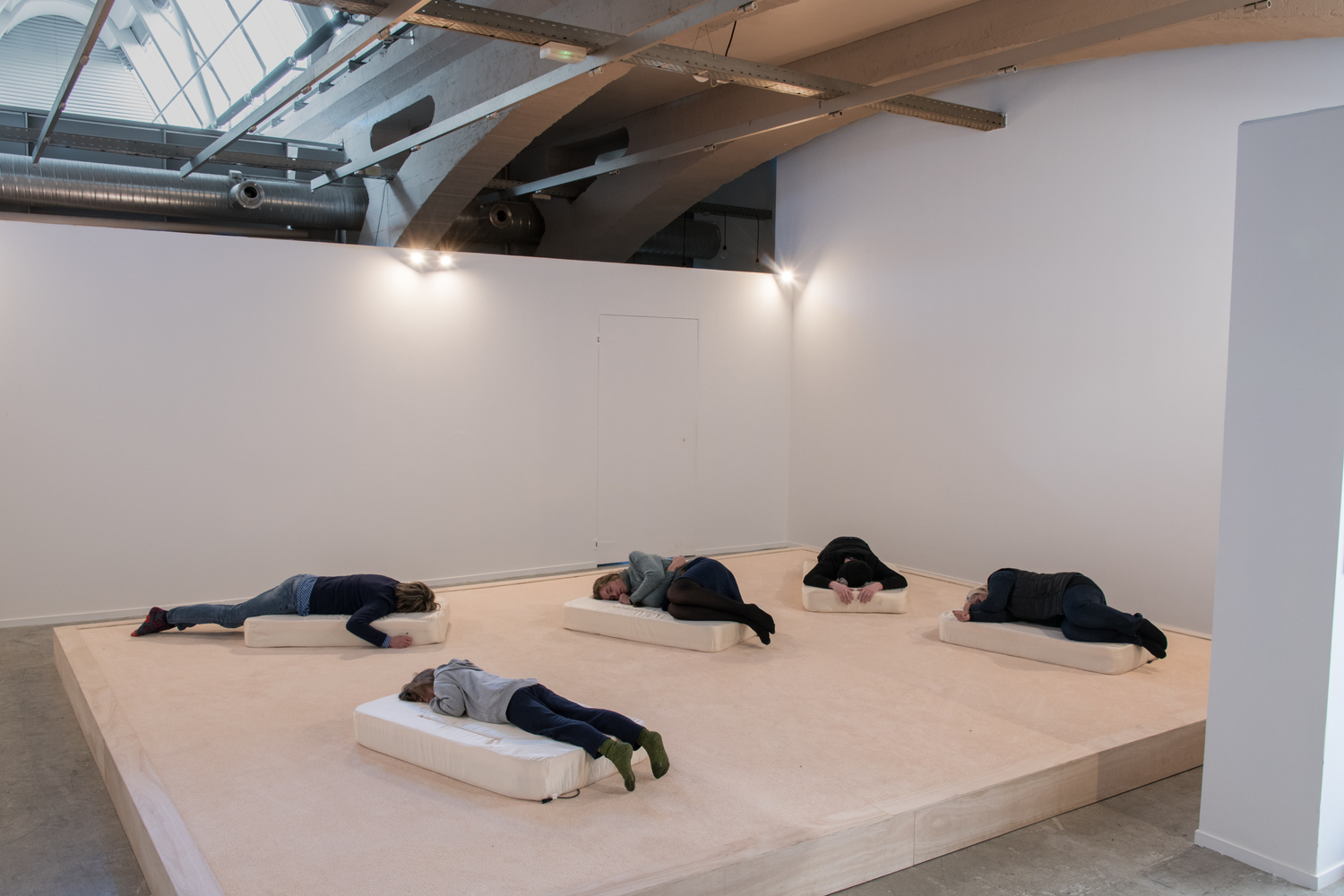 documentation image of the installation. there are five people laying on their respective mattresses , two are laying sideways and the other three are facing down. the mattresses are on a large block of plywood creating a separation between the installation and the floor, there aren't any curtains in this installation.