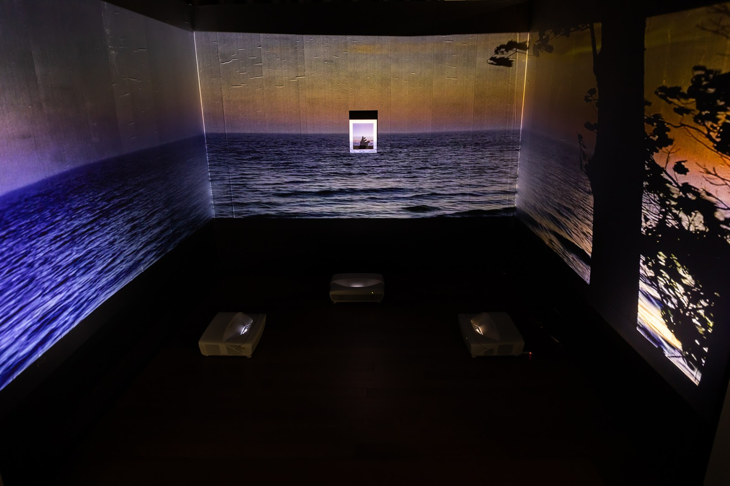 A dimly lit room-sized art installation where a projection of a sunset seascape of dark blues, purples, and oranges, covers three sides of a closed in space. In the very middle of the center wall is a small framed photograph depicting, perhaps, a small figure against a sky. 