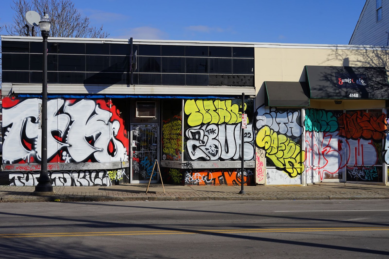 An exterior view of a storefront type building on a road and sidewalk that has been covered in graffiti tags that has been tightly packed together. 