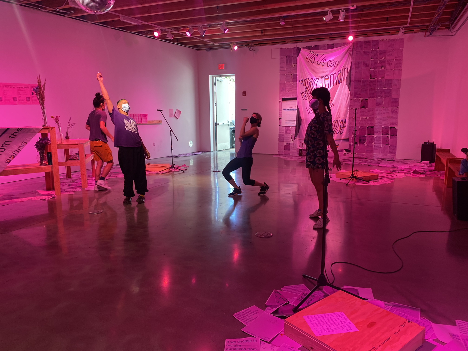 people standing in a pink exhibition space gesture in different ways with their bodies and arms. one person, on the left side, raises their fist and toroso toward the cieling, another person begins to kneal and bends their elbows, while another stretches their limbs toward their sides.