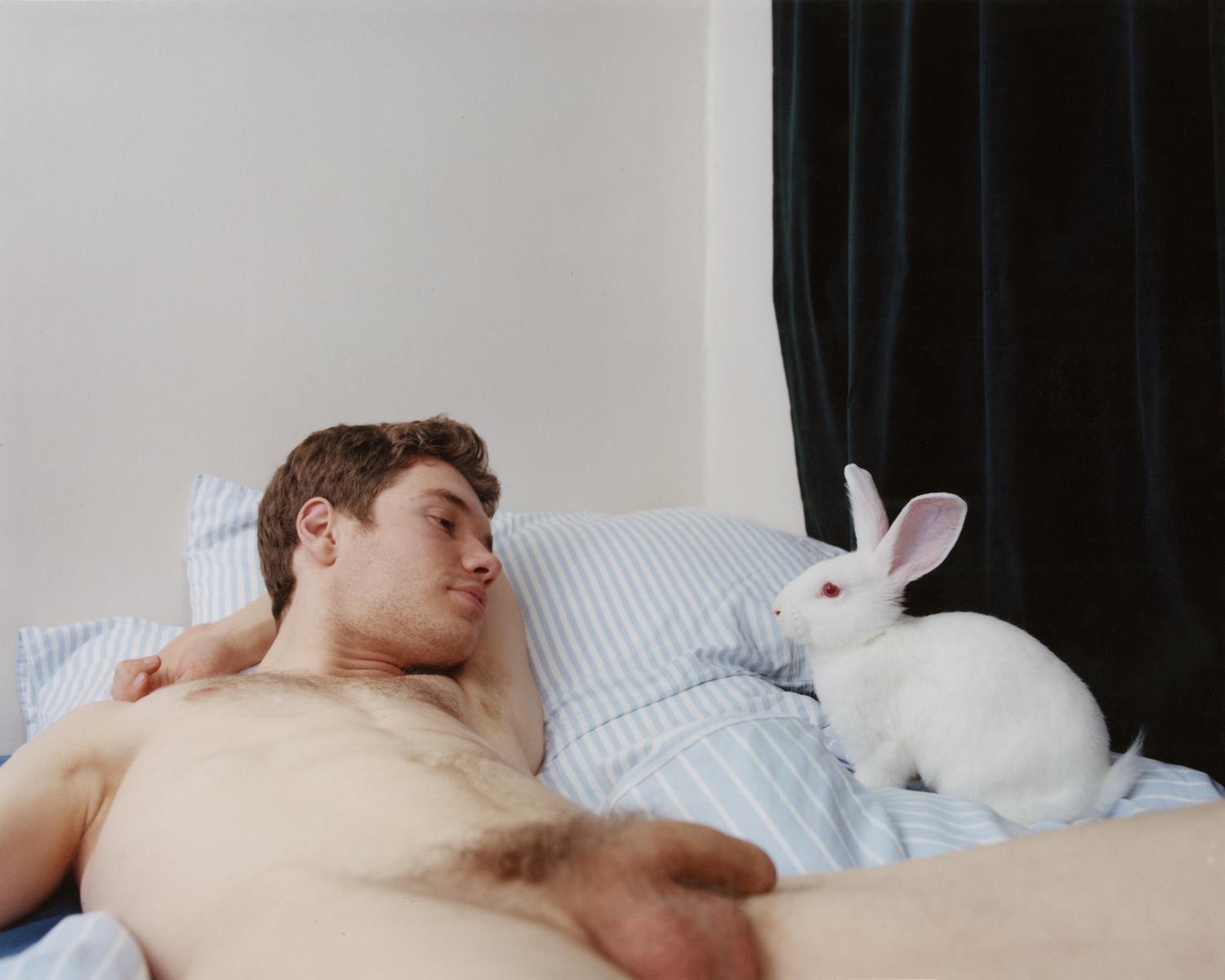 Un jeune homme blanc allongé sur un lit face à un lapin blanc