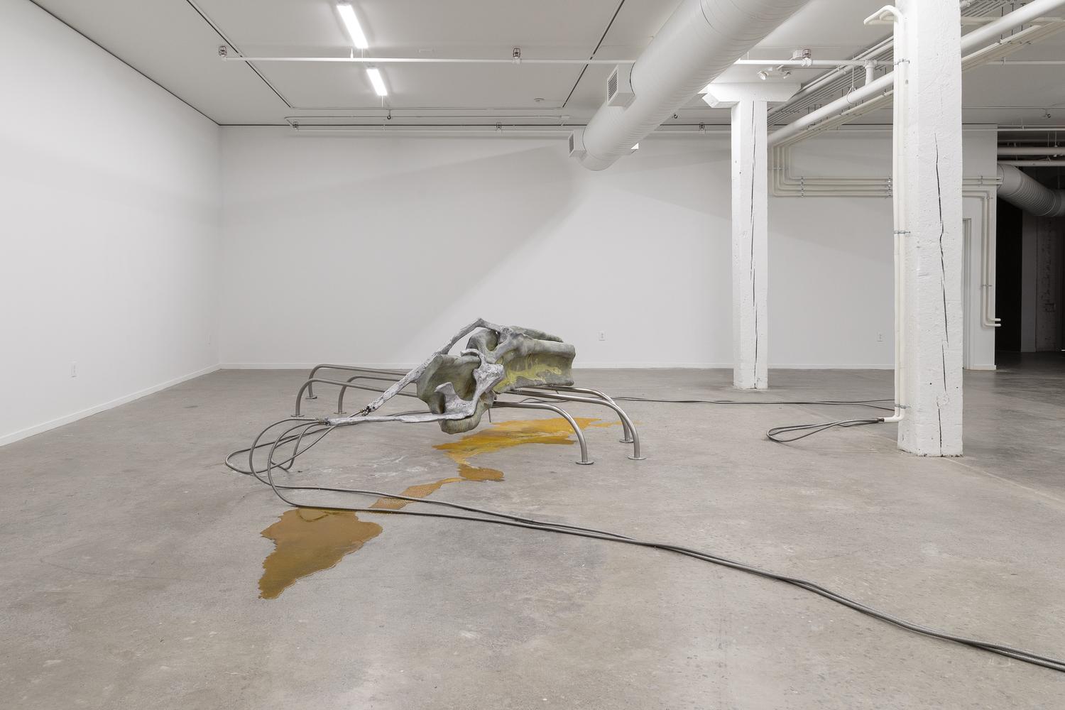 An art gallery with white walls and concrete floors, which is empty save for an abstract metal sculpture that resembles something between a spider and a pile of dinosaur bones. It seems to be leaking a dark orange hued fluid, like car engine oil.