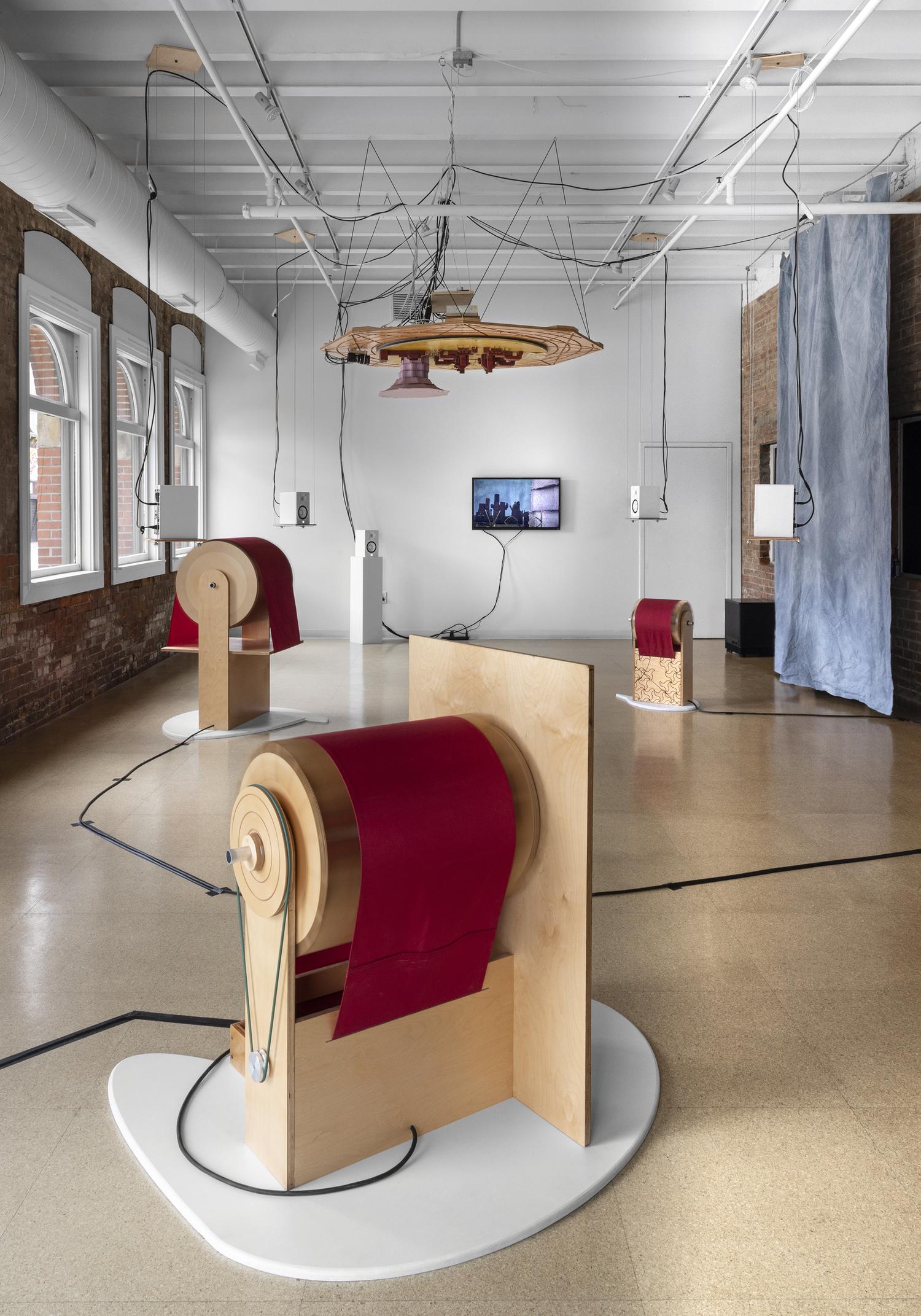 A gallery space with several floor standing sculptures resembling motors or barrels that seem to all be electrically powered with many black wires connecting them all.