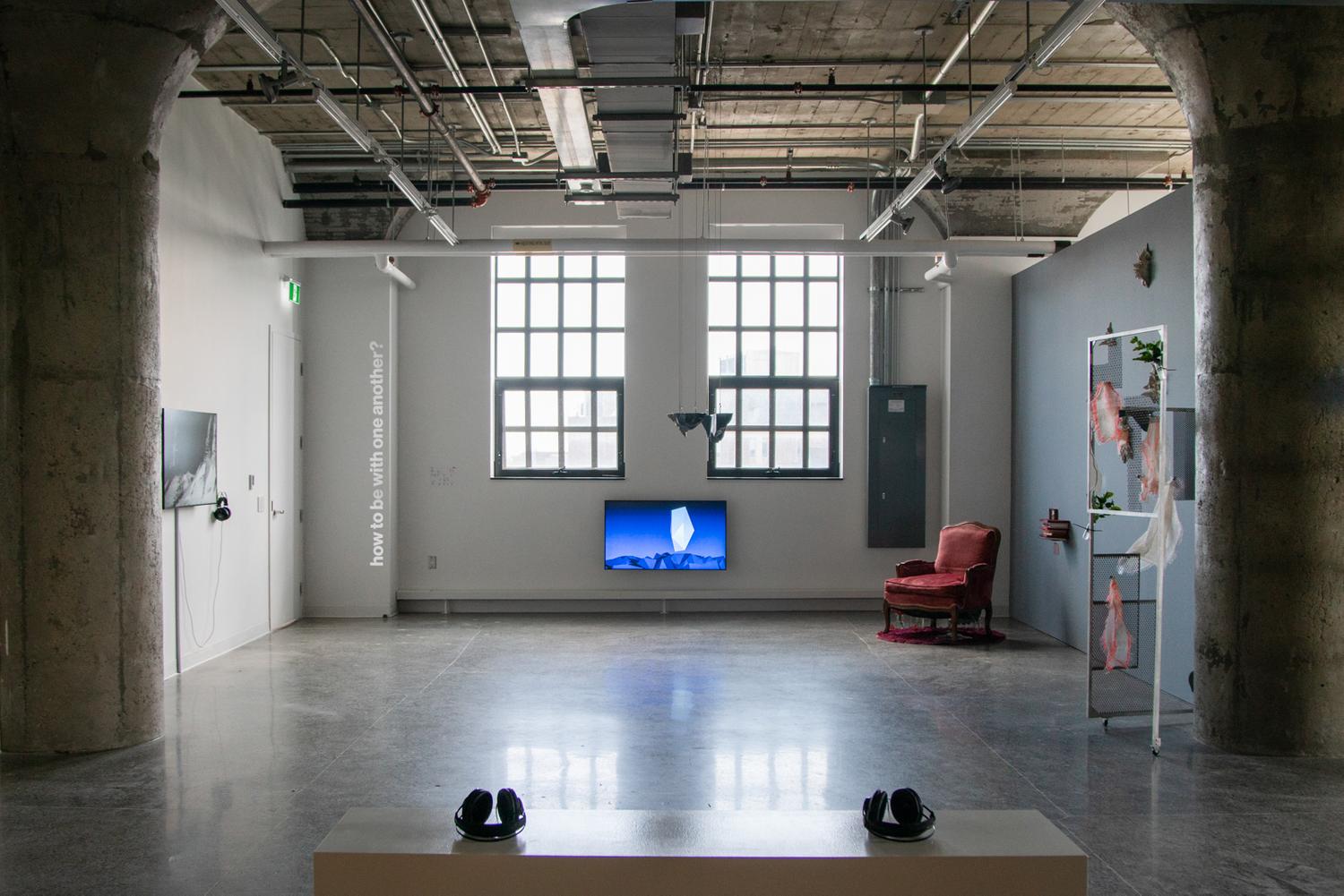 A photo of the south side of the gallery, taken from in the middle of a  gallery. In the foreground, two black headsets rest on a white bench in the centre of the room. An audio piece by Workman. Behind it, various artworks are displayed along the three walls of the gallery. Viewed from left to right in the photo: a TV is hung on eye-level, screening a black-and-white video work by Kolcze. On the white wall, a silvery vinyl text reads, quote, how to be with one another?, end quote. A TV monitor hangs low on the wall of the gallery centred between and below two big windows, screening an animated video by Nuff of a crystal-like landscape. A small black sculpture by Polymetis hangs from the ceiling, in the middle of the room. The wall on the right is dark grey. Against it, a red velvet chair and a stack of zines by Woudenberg. Grey sculptures scattered on the wall by Salazar. A sculptural installation featuring an aluminum door frame, bandages, and plants by Dreifelds.