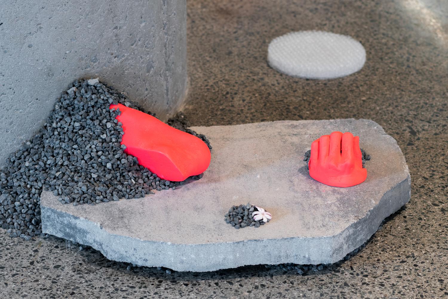 Close-up of neon orange assemblage by Dreifelds, showing partial casts of a hand and a foot in neon resting on a slab of stone and gravel with a jasmine flower. A stack of bubblewrap on the background.