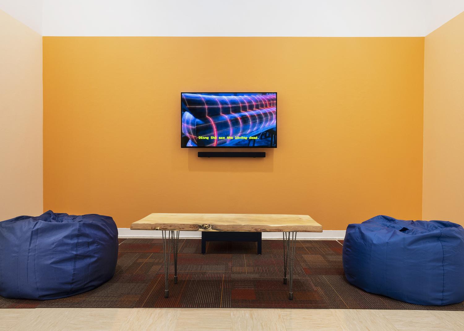 A TV is mounted on a yellow wall showing an ambiguous image of blue and pink shapes with subtitles that read “Jiang Chi are the living dead.” In front of the TV is a wooden table with a live edge finish, flanked on each side by two oversized blue bean bag chairs.