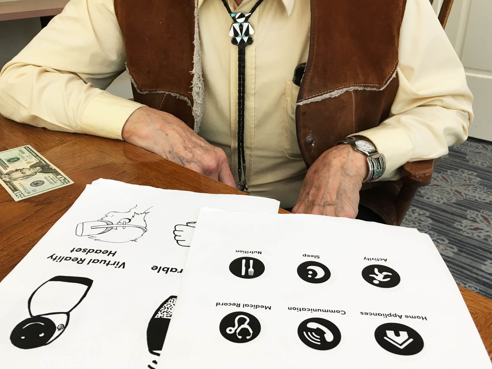 The torso of a man sitting in a chair. In front of him on a table are illustrated pages of technologies and uses.