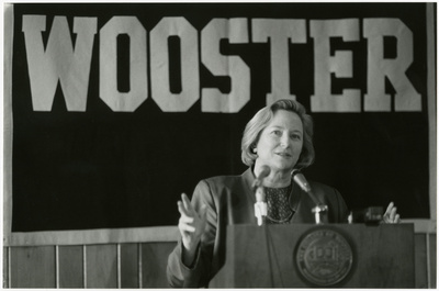 A black and white press conference photograph of Susanne Woods standing behind a podium, with a banner that reads “WOOSTER” hung behind her, and the college’s seal on the front of the pedestal.