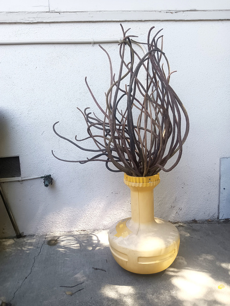A sculpture in resemblance of a flame sitting on the ground outdoors and in front of a stucco wall.