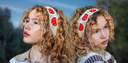A headshot of two human figures that look identical to each other, are dressed in the same way, and are posing back to back, each looking toward something out of the frame. By the way their hair is touching, they seem almost contiguous to each other. Natural landscape in the background is blurred. 