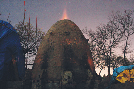 A mud house with fire from within is installed outdoors in nature.