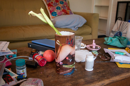 A cylinder vase with photo transfer of a nude atop a coffee table with two stems of calla lilies inserted into their anus holds two stems of calla lilies. The vase is placed on the same coffee table cluttered with mundane objects including mails, pills, a ball, a pair of glasses, a pile of a journal and printed materials, masks, a candle, antibacterial wipes, and more.