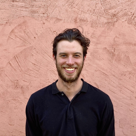 A person with a big smile gazing directly forward. They have short wavy brown hair with a beard and mustache. They are wearing a dark blue collared long sleeve shirt. Behind them is a terra cotta wall. 