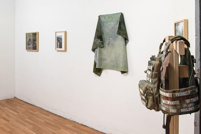 A white wall displaying four artworks with a sculpture in the foreground. From left to right, there are two photos in light wood frames on the wall, followed by a photograph printed on fabric and draped on the wall, and then another framed photo. The sculpture is a plate carrier vest with patches embroidered with white text, resting on a wood stand.
