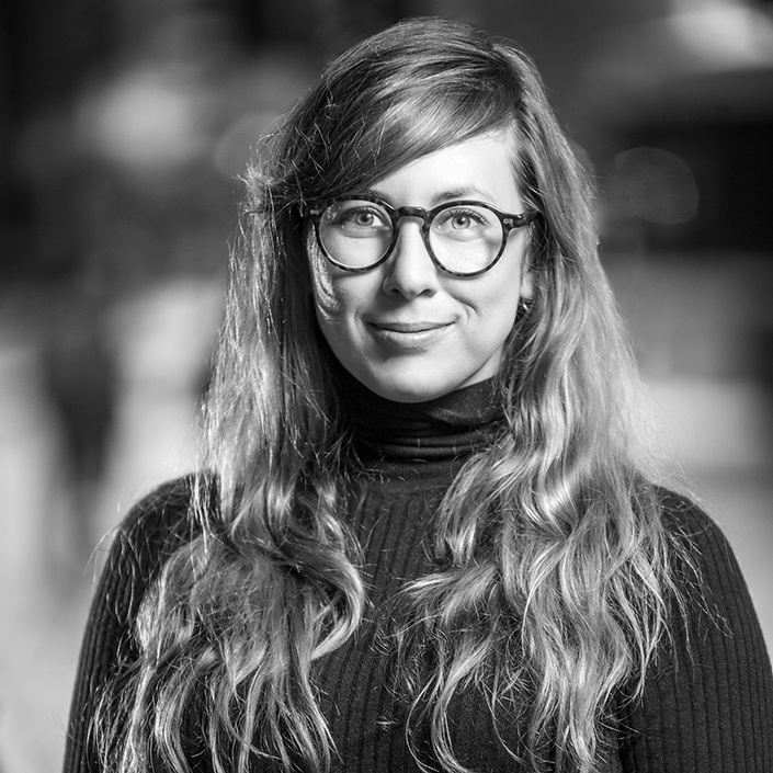 Person with long hair wearing a turtleneck with thick black glasses staring straight ahead and smiling.