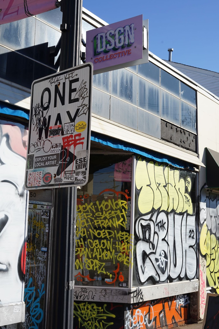 A “One Way” traffic sign that has been covered in graffiti in front of an angled building front that also been covered in graffiti. Above is a sign that reads “DSGN Collective”