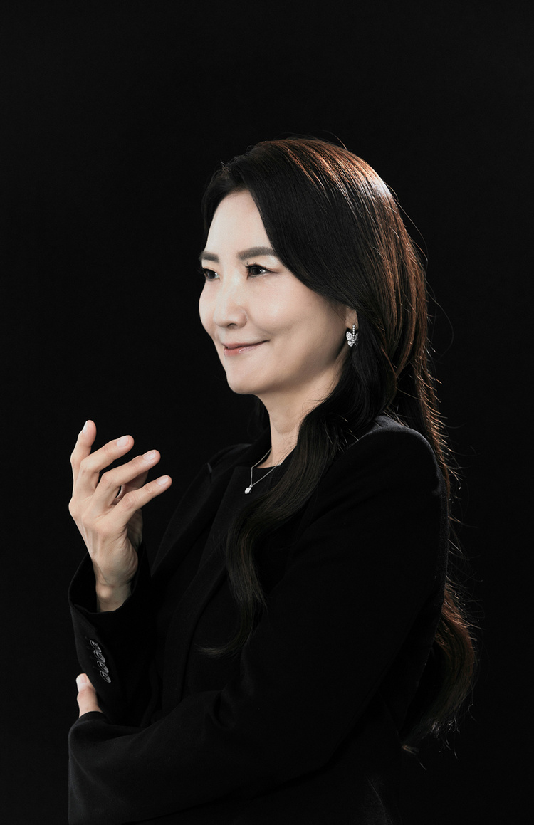 A half body profile of pianist Jenny Soonjin Kim. She looks ahead with a slight smile. Her arms are folded; and the right hand is raised to the chest height, relaxed with the palm backward.