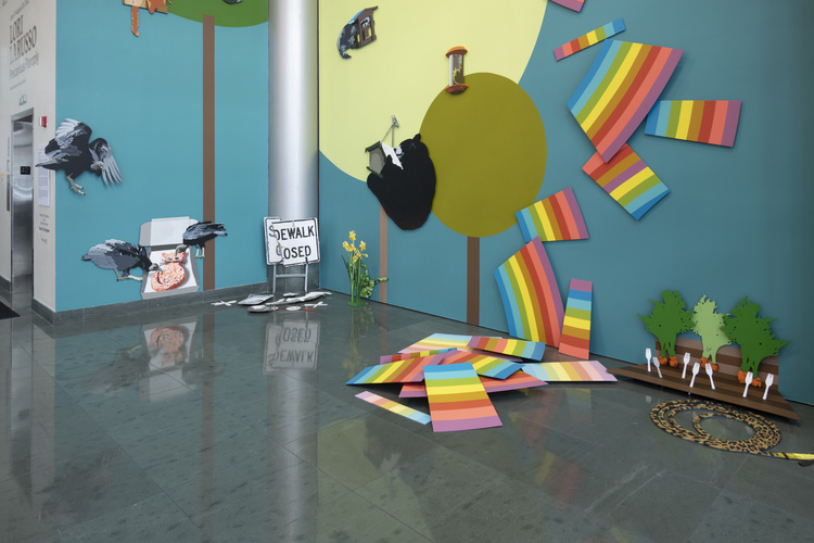 Various painted flat panels of many different shapes come together to form a surreal outdoor scene that includes a rainbow falling to the ground in many pieces in a pile on the floor, surrounded by a small garden of carrots, round trees, vultures eating a to-go pizza from the box, and a bent sign that reads “sidewalk closed.”