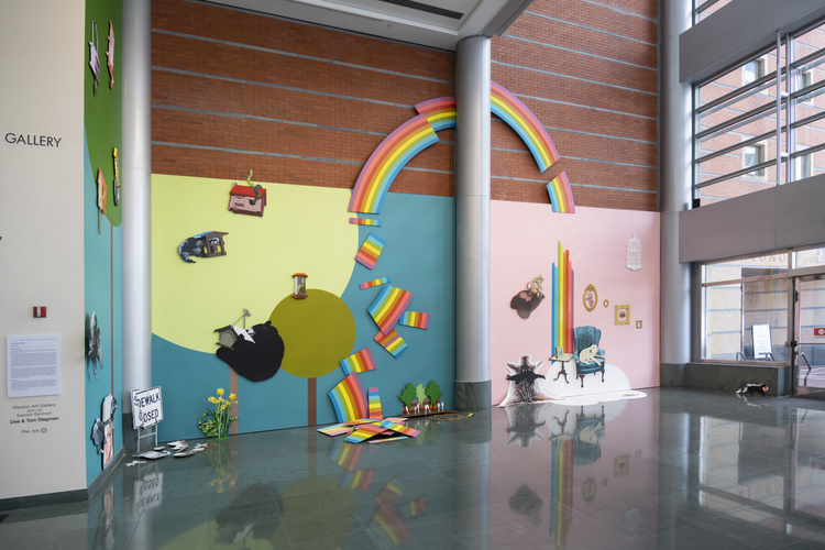 Wide angle view of a colorful art installation that has many differently painted background walls behind flattened painted images of things like a broken up rainbow, a surreal living room scene, trees and a sky.