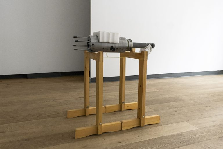 A photo of the heavy steel part known as a plunger/barrel assembly that came from the Atlantic Conveyor. It is sitting on a wooden stand in the gallery. 
