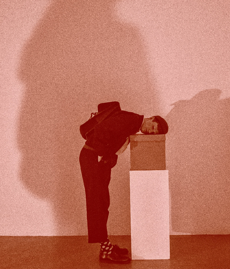 Calum Perrin, a young man wearing a dark jacket and cropped trousers, rests their head on a cardboard box placed above a plinth.