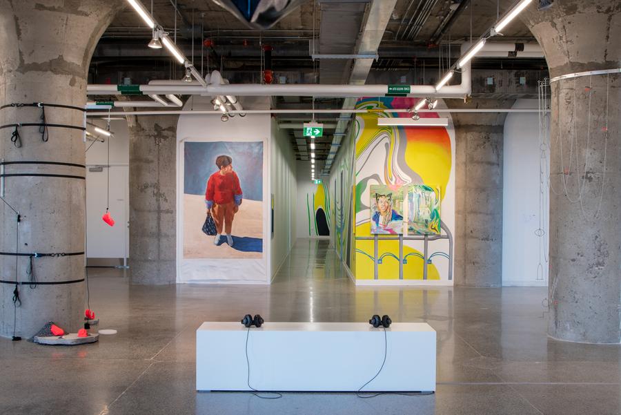 A photo of the north side of the gallery. Workman's audio piece on the white bench again in the centre foreground. Behind it, depicted from left to right, Dreifelds's black-straps, neon-sculpture assemblage on the grey column. Olou's painting of a figure in a professional and stylish clothing, holding a lunch bag. A mural and tryptich by McComas goes down a long hallway, depicting a figure seemingly in exploration of a dream-world construction site with aluminum beams. Dreifeld's assemblage of chainlink drapings on another grey structural column.